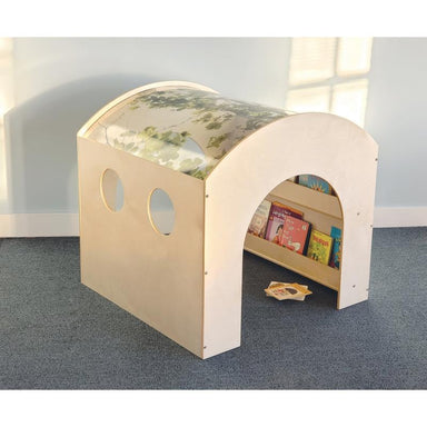 A children's wooden playhouse or reading nook with an arched opening, circular window cutouts, and a small shelf inside holding colorful books. The structure is shown in a room with blue carpet and a pale wall.