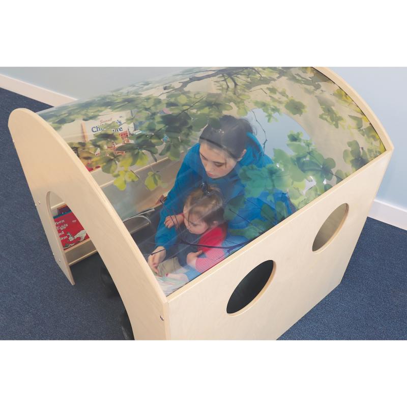 A wooden children's play tunnel with a printed green forest canopy overhead, shown in a classroom setting with children and an adult inside.