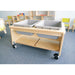 A light wood toddler activity table with two gray plastic bins on top and two lower storage shelves, shown in a classroom setting on a blue carpet.